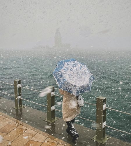 A person holding a blue umbrella walking by a snowy waterfront, with a distant view of a lighthouse.